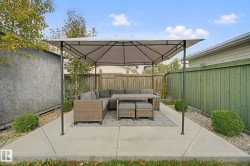Paved patio area with a covered gazebo structure, bordered by a privacy fence and green shrubbery - 