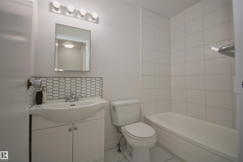 White subway tile shower surround, integrated bathtub, white vanity with storage, mosaic tile backsplash, and wall-mounted vanity lighting - 7743 37 Avenue Nw, Edmonton, AB 
