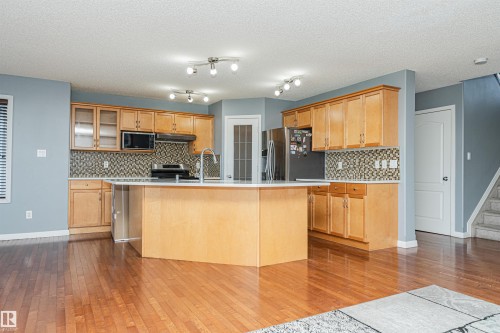Spacious kitchen featuring wood-finish cabinetry, a large central island with an integrated sink, mosaic tile backsplash, and stainless steel appliances - 1254 Mcallister Way Sw, Edmonton, AB 