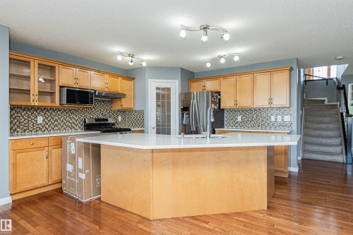Kitchen featuring a large island with a light-toned countertop, wood-finish cabinetry, stainless steel appliances, and a mosaic tile backsplash - 1254 Mcallister Way Sw, Edmonton, AB 