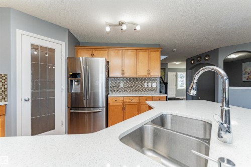 Kitchen featuring a double basin sink, stainless steel appliances, light wood-finish cabinetry, mosaic tile backsplash, and light-colored countertops - 1254 Mcallister Way Sw, Edmonton, AB 