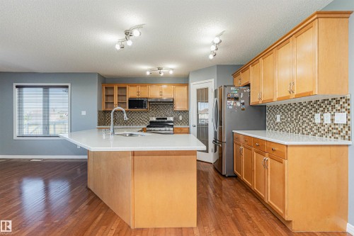 Kitchen featuring wood-finish flooring, light wood cabinetry, a mosaic tile backsplash, stainless steel appliances, and a central island with a sink - 1254 Mcallister Way Sw, Edmonton, AB 