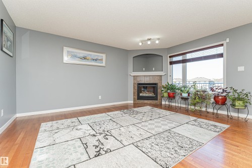 Hardwood-style flooring, a corner fireplace with a tiled surround, a large window, a built-in display alcove, and track lighting - 1254 Mcallister Way Sw, Edmonton, AB 