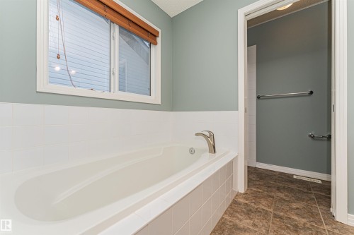 Built-in bathtub with a white tiled surround, window with wood-finish blinds, and a brushed nickel faucet - 1254 Mcallister Way Sw, Edmonton, AB 