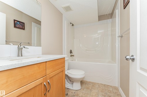Bathroom featuring a single vanity with wood-finish cabinetry, a light-toned countertop, a white subway tile backsplash, and a tub-shower combination with a white surround - 1254 Mcallister Way Sw, Edmonton, AB 