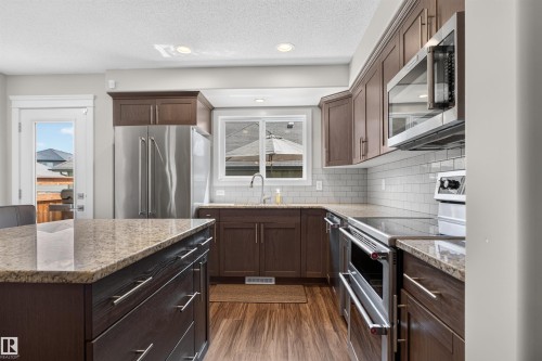 Contemporary kitchen featuring dark wood cabinetry, stainless steel appliances, speckled stone countertops, and wood-finish flooring - 319 Desrochers Boulevard Sw, Edmonton, AB 