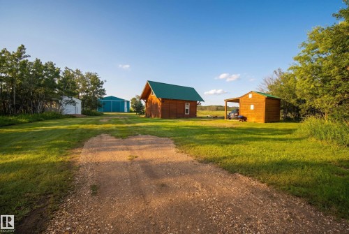 Gravel drive leading to multiple outbuildings, including a blue metal barn, a wood-sided structure with a green metal roof, and an open-sided wood carport - 48109 Rr 155, Rural Beaver County, AB - Outdoor