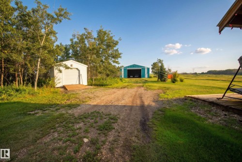 Expansive outdoor space featuring a detached white storage shed with a ramp, a large teal outbuilding with an open bay, mature trees, and a wooden deck - 48109 Rr 155, Rural Beaver County, AB - Outdoor