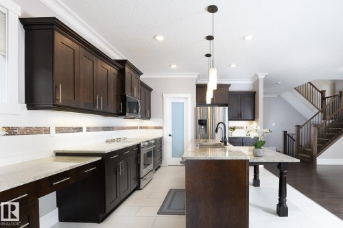 Kitchen featuring dark wood cabinetry, light-toned countertops, stainless steel appliances, and an expansive island with a sink and pendant lighting - 886 Armitage Wynd Sw, Edmonton, AB 