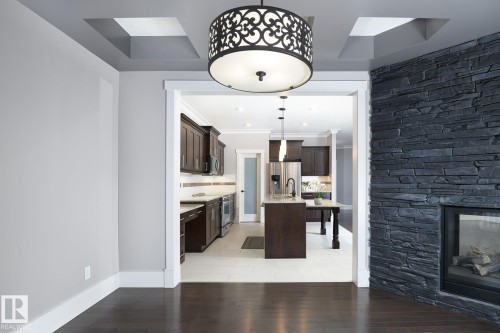 Hardwood-finish flooring, recessed ceiling with decorative lighting, stone-clad fireplace, kitchen with dark wood-finish cabinetry, and stainless steel appliances - 886 Armitage Wynd Sw, Edmonton, AB 