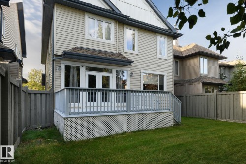 Rear exterior featuring light-colored siding, a wooden deck with railing, dual glass-paneled doors, and a spacious green lawn - 886 Armitage Wynd Sw, Edmonton, AB 