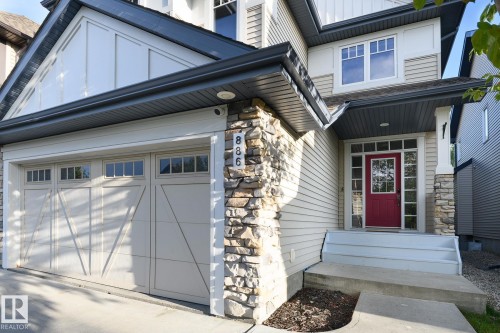 Stone accent facade with siding, two-car garage with transom windows, covered entry with a red door, and multiple windows - 886 Armitage Wynd Sw, Edmonton, AB 