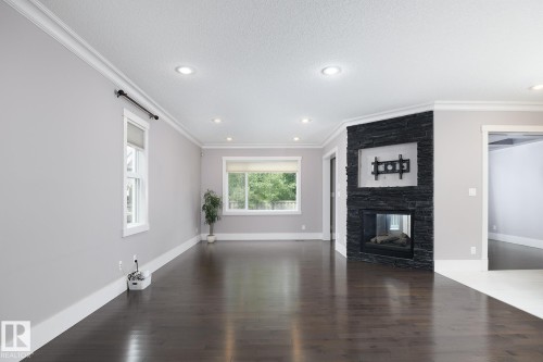 Spacious interior featuring dark wood-finish flooring, a stone-clad fireplace, recessed lighting, white crown molding, and large windows - 886 Armitage Wynd Sw, Edmonton, AB 