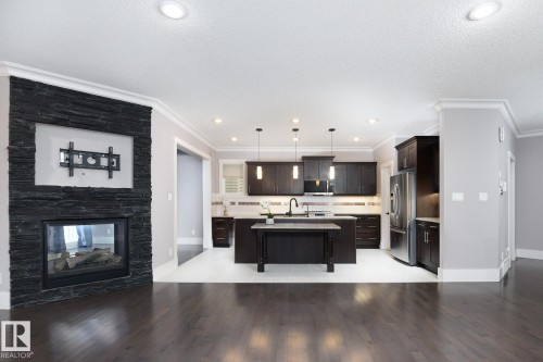 Dark wood-finish flooring, a contemporary fireplace with a stone-clad surround, recessed ceiling lighting, a kitchen island, and dark wood cabinetry with integrated appliances - 886 Armitage Wynd Sw, Edmonton, AB 