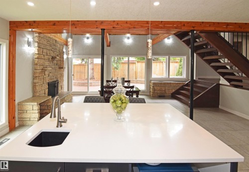 Quartz countertop island with an undermount sink, a stone-clad fireplace, exposed wood beams, a custom wooden staircase with metal railings, and a sliding glass door to the exterior - 9657 68A Street Nw, Edmonton, AB 