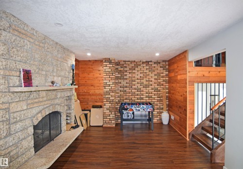 Stone-clad fireplace with hearth, wood-finish flooring, accent brick wall, wood paneling, and a staircase with metal railings - 9657 68A Street Nw, Edmonton, AB 