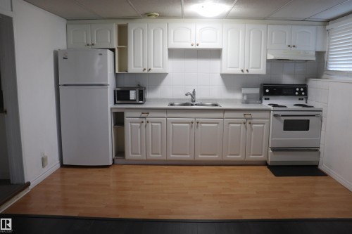 Kitchen featuring white shaker-style cabinetry, a white tile backsplash, light-toned wood-finish flooring, a stainless steel sink, and white appliances - 8828 151 Avenue Northwest, Edmonton, AB 