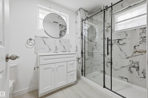 Bathroom featuring a white vanity with integrated sink, a circular hanging mirror, a glass-enclosed shower with black framing, and marble-patterned wall tiling - 13024 Sherbrooke Avenue Nw, Edmonton, AB 