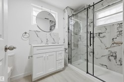 Bathroom featuring a white vanity with integrated sink, a circular hanging mirror, a glass-enclosed shower with black framing, and marble-patterned wall tiling - 