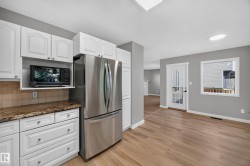 White cabinetry with brushed nickel hardware, stone-look countertops with a tile backsplash, stainless steel refrigerator, light wood-finish flooring, and a glass-paneled door - 