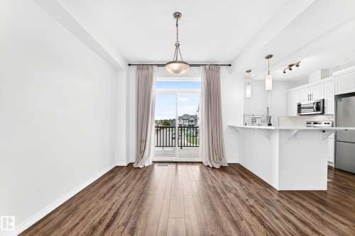 Open-concept living space featuring wood-finish flooring, a sliding glass door with balcony access, and a kitchen with white cabinetry and stainless steel appliances - 20008 19 Avenue Nw, Edmonton, AB 