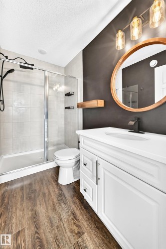 Wood-finish flooring, white vanity with integrated sink, glass-enclosed shower with tile surround, wood-framed mirror, and modern vanity lighting - 20008 19 Avenue Nw, Edmonton, AB 
