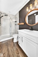 Wood-finish flooring, white vanity with integrated sink, glass-enclosed shower with tile surround, wood-framed mirror, and modern vanity lighting - 