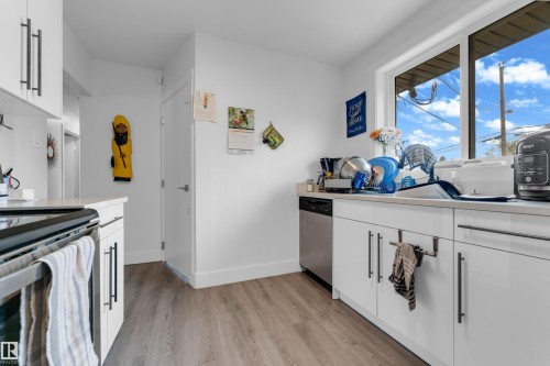 Kitchen featuring wood-finish flooring, white cabinetry with dark hardware, stainless steel dishwasher, and a window with a white frame - 14216 58 Street Nw, Edmonton, AB 