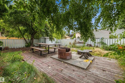 Expansive outdoor decking with a central fire pit, surrounded by mature trees, a white picket fence, and a utility shed - 10621 60A Avenue Nw, Edmonton, AB 