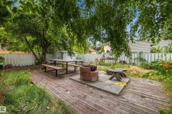 Expansive outdoor decking with a central fire pit, surrounded by mature trees, a white picket fence, and a utility shed - 