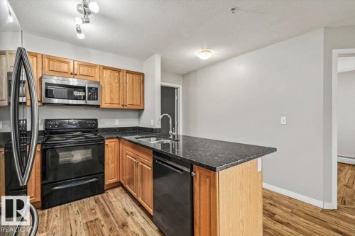 Kitchen featuring wood-finish flooring, dark stone countertops, wood cabinetry, stainless steel microwave, and black range - #110 4309 33 St, Stony Plain, AB 