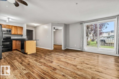 Expansive main living area featuring wood-finish flooring, a kitchen with wood cabinetry and black appliances, an island with a sink, and a large sliding glass door - #110 4309 33 St, Stony Plain, AB 