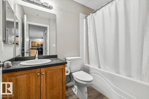 Bathroom featuring a wood-finish vanity with an integrated sink, a dark countertop, an expansive mirror, and a white bathtub with tile surround - #110 4309 33 St, Stony Plain, AB 