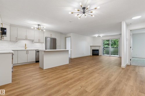 Open-concept living space featuring wood-finish flooring, a contemporary kitchen with light-colored cabinetry and an island, stainless steel appliances, a tile backsplash, and a fireplace with a mantel - #353 2096 Blackmud Creek Dr Sw, Edmonton, AB - Indoor Photo Showing Kitchen With Upgraded Kitchen