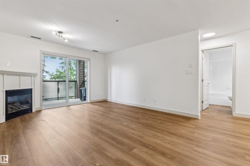 Open-concept living area featuring wood-finish flooring, a contemporary fireplace with a white mantel, and a sliding glass door opening to a private balcony - #353 2096 Blackmud Creek Dr Sw, Edmonton, AB - Indoor Photo Showing Living Room With Fireplace