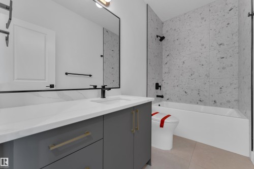 Modern bathroom featuring a single vanity with a white countertop and gray cabinetry, a black framed mirror, and a bathtub with stone-look wall tiling - 1129 Starling Drive Nw, Edmonton, AB 
