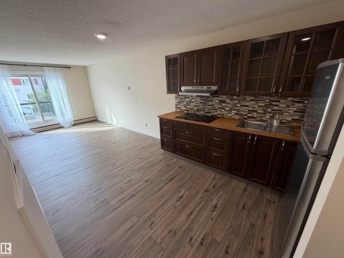 Open-concept living area with wood-finish flooring, a full kitchen featuring dark wood cabinetry, a mosaic tile backsplash, and a stainless steel refrigerator - #209 11429 124 St Nw, Edmonton, AB 