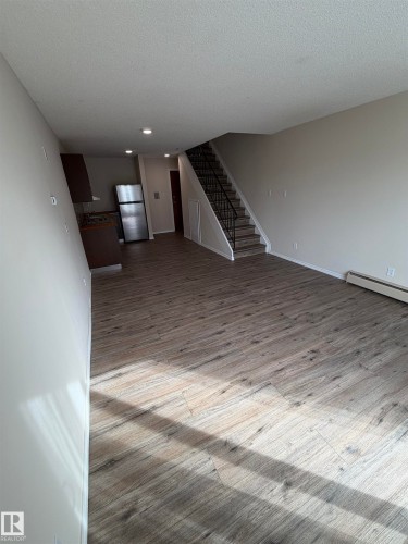 Wood-finish flooring throughout, stainless steel refrigerator, dark wood-finish cabinetry, recessed ceiling lighting, and a staircase with an ornamental metal railing - #209 11429 124 St Nw, Edmonton, AB 