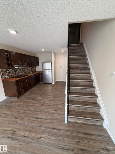 Open-concept living area with wood-finish flooring, recessed lighting, a fully equipped kitchen featuring dark wood cabinetry, and a staircase with a black metal handrail - #209 11429 124 St Nw, Edmonton, AB 