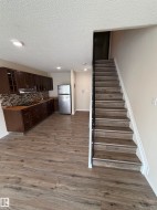 Open-concept living area with wood-finish flooring, recessed lighting, a fully equipped kitchen featuring dark wood cabinetry, and a staircase with a black metal handrail - 