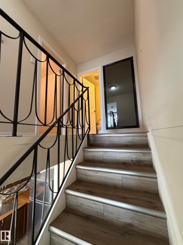 Staircase featuring wood-finish treads with metal trim, decorative wrought iron railing, and a full-length wall mirror - #209 11429 124 St Nw, Edmonton, AB 