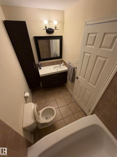 Full bathroom featuring a single vanity with storage, framed mirror, wall sconce lighting, and a white paneled door - #209 11429 124 St Nw, Edmonton, AB 