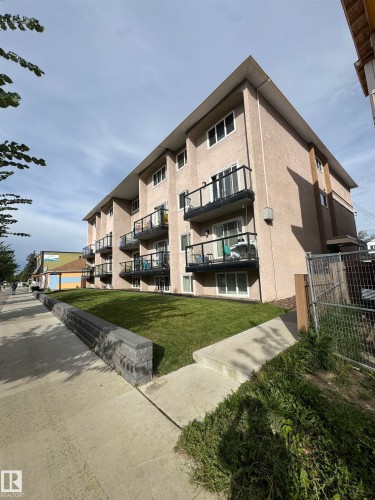 Multi-story building featuring stucco exterior, multiple balconies with glass railings, and a sloped lawn with a retaining wall - #209 11429 124 St Nw, Edmonton, AB 