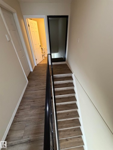 Wood-finish flooring throughout the hallway and stair treads, featuring a contrasting black metal handrail and white trim - #209 11429 124 St Nw, Edmonton, AB 