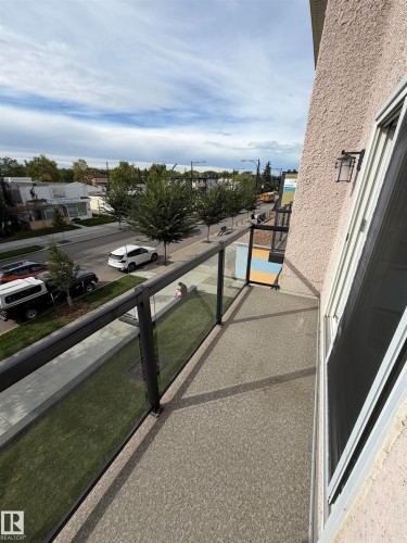 Private balcony featuring textured flooring, clear glass railings with dark frames, and an exterior wall-mounted light fixture - #209 11429 124 St Nw, Edmonton, AB 