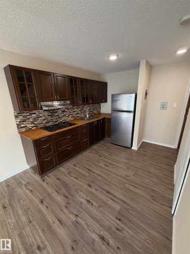 Kitchen featuring dark wood-finish cabinetry, a tiled mosaic backsplash, wood-finish countertops, stainless steel appliances, and wood-finish flooring - #209 11429 124 St Nw, Edmonton, AB 