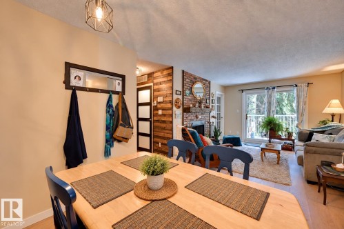 Open concept living area featuring a light-colored wood-finish table, a decorative geometric pendant light, a textured wood accent wall, a brick fireplace, and a sliding glass door with balcony access - #203 24 Alpine Pl, St. Albert, AB 