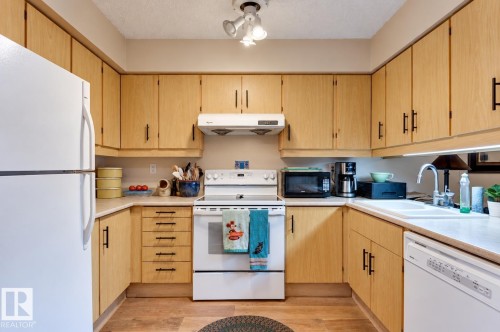 U-shaped kitchen featuring wood-finish cabinetry, white appliances, a white sink, and wood-finish flooring - #203 24 Alpine Pl, St. Albert, AB 
