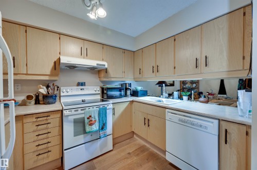 Wood-finish flooring, light wood cabinetry with dark hardware, white countertop surfaces, a built-in white dishwasher, and a white electric range with an overhead vent - #203 24 Alpine Pl, St. Albert, AB 