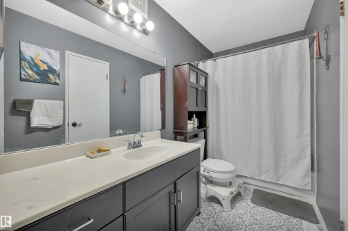 Bathroom featuring a dark gray vanity with a single basin sink, wall-mounted light fixture, full-width mirror, and patterned floor tiles - 15017 26 Street Nw, Edmonton, AB 
